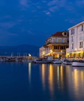 Waterfront scene featuring boats docked near a building with lights reflecting on the water at dusk. - Olive Oil Times