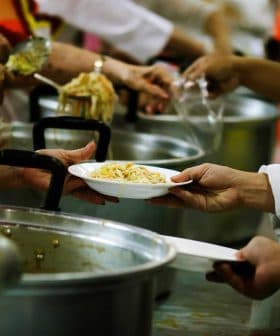 Hands serving a plate of food from a large pot in a community kitchen setting. - Olive Oil Times