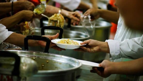 Hands serving a plate of food from a large pot in a community kitchen setting. - Olive Oil Times