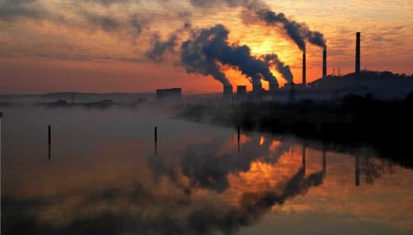 Silhouetted industrial power plant with smoke stacks against a sunset sky and water reflection. - Olive Oil Times