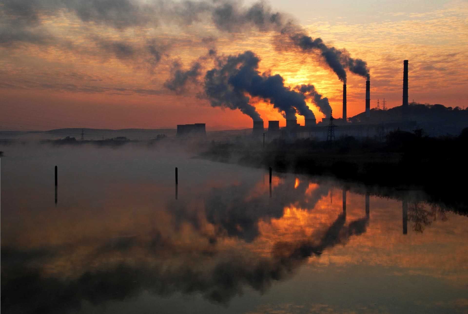 Silhouetted industrial power plant with smoke stacks against a sunset sky and water reflection. - Olive Oil Times