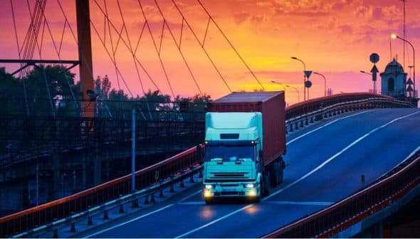 A white truck with a container driving on a bridge during sunset with colorful sky. - Olive Oil Times