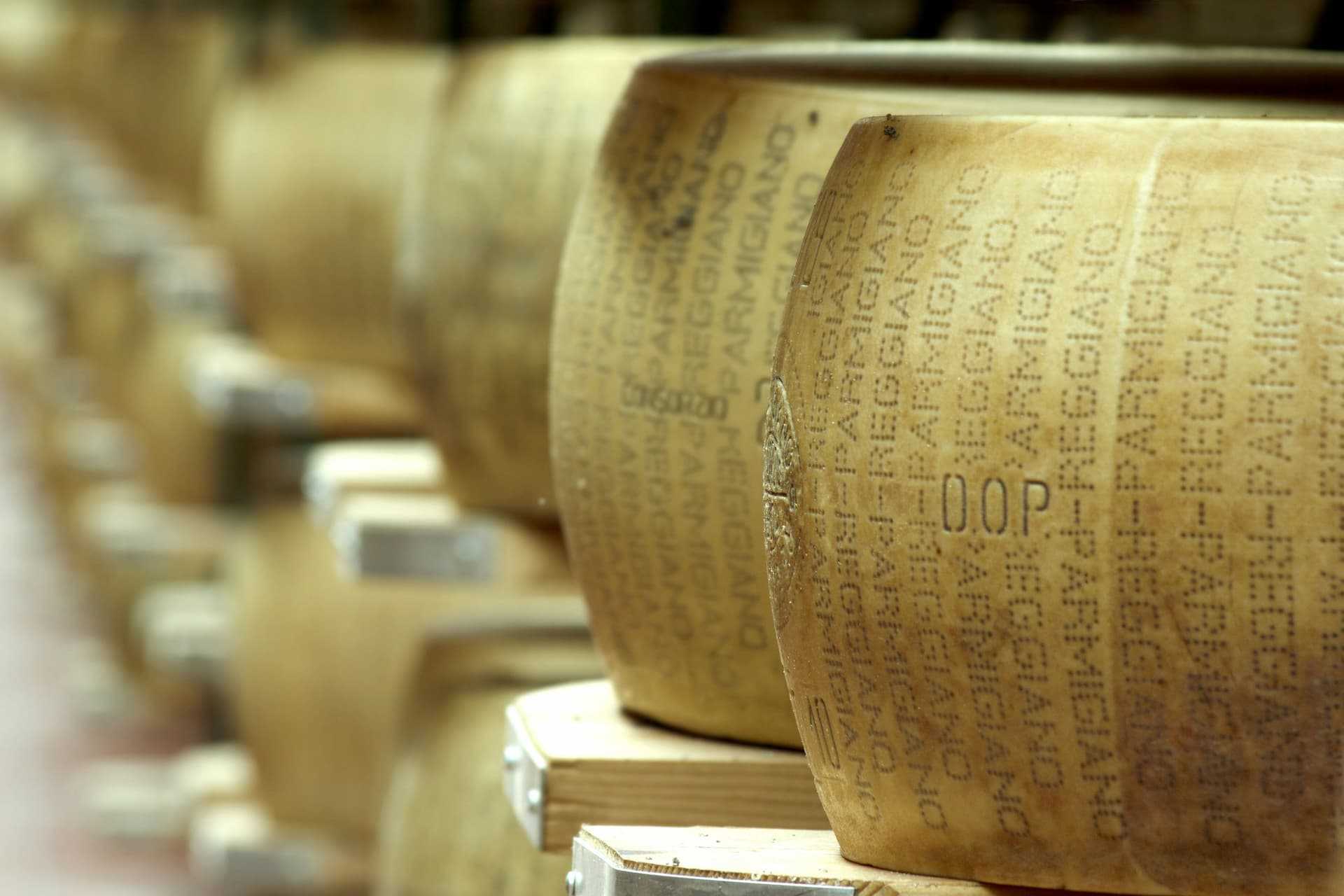 Close-up of aged cheese wheels with embossed labels stacked on wooden shelves. - Olive Oil Times