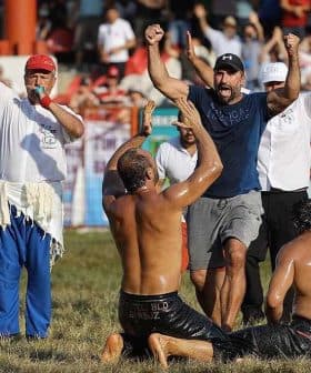 Two men wrestling on the ground while a referee and spectators cheer in the background. - Olive Oil Times