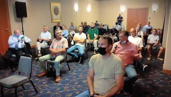 Group of people seated in a meeting room, some wearing masks, listening to a speaker. - Olive Oil Times