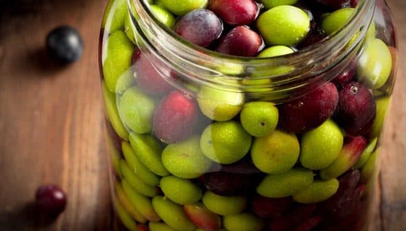 Glass jar filled with a mix of green and black olives on a wooden surface. - Olive Oil Times