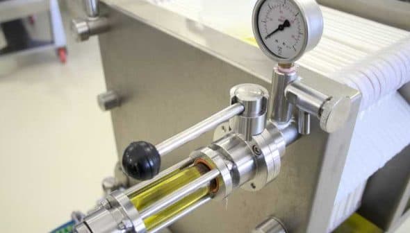 Close-up of oil filtration equipment showing a pressure gauge and transparent tube with liquid. - Olive Oil Times
