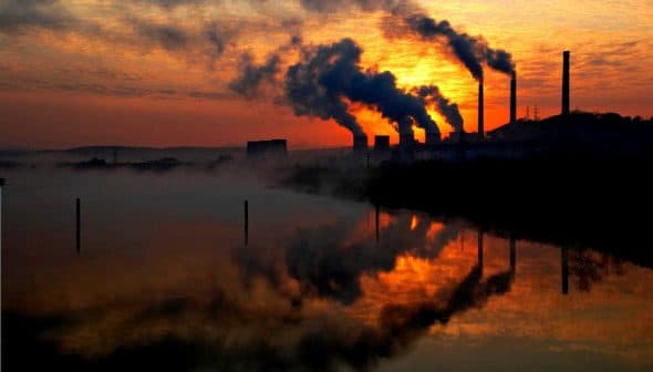 Silhouetted industrial smokestacks emitting smoke against a sunset sky with reflections in water. - Olive Oil Times