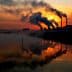 Silhouetted industrial smokestacks emitting smoke against a sunset sky with reflections in water. - Olive Oil Times
