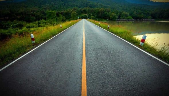 A straight road lined with white and red markers, leading through a green landscape with mountains in the background. - Olive Oil Times