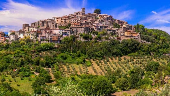 A hilltop village with stone buildings and olive groves in the foreground under a blue sky. - Olive Oil Times