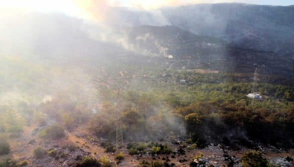 Aerial view of a landscape with smoke rising from the ground among trees and rocky terrain. - Olive Oil Times