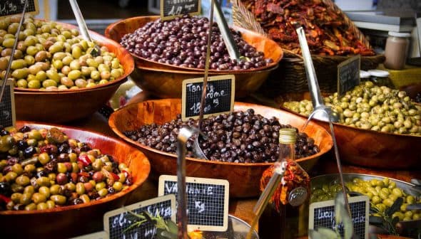 Assorted olives displayed in wooden bowls at a market, with labels indicating prices. - Olive Oil Times