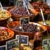 Assorted olives displayed in wooden bowls at a market, with labels indicating prices. - Olive Oil Times