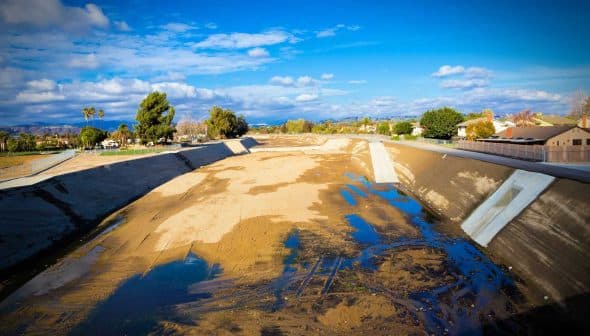 A dry riverbed with patches of water and sparse vegetation under a blue sky with clouds. - Olive Oil Times