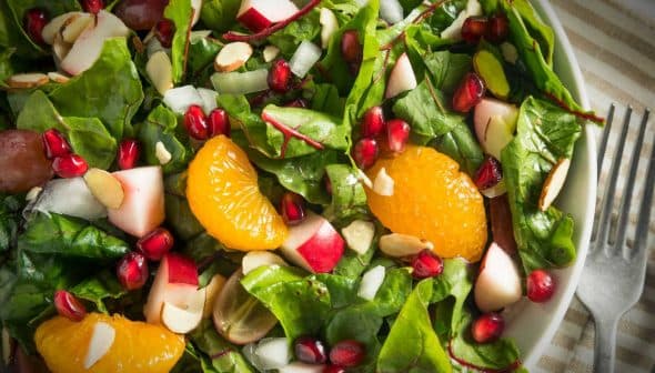 A bowl of mixed green salad featuring spinach, apple pieces, orange segments, pomegranate seeds, and sliced almonds. - Olive Oil Times