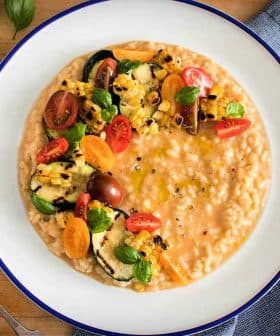Bowl of creamy risotto topped with grilled vegetables and cherry tomatoes on a wooden table. - Olive Oil Times