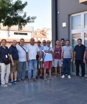 A group of individuals standing together in a line outside a building, some wearing sunglasses and casual clothing. - Olive Oil Times