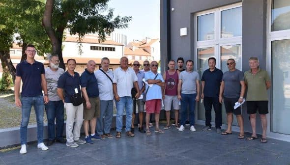 A group of individuals standing together in a line outside a building, some wearing sunglasses and casual clothing. - Olive Oil Times