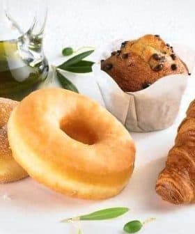 Two sugar-coated donuts, a chocolate chip muffin, and a croissant on a plate with a glass of olive oil. - Olive Oil Times