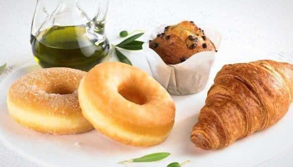 Two sugar-coated donuts, a chocolate chip muffin, and a croissant on a plate with a glass of olive oil. - Olive Oil Times
