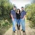 Three individuals standing together in an olive grove, wearing casual clothing and hats. - Olive Oil Times