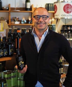 Man with glasses holding a bottle of olive oil in a store filled with various oil products. - Olive Oil Times