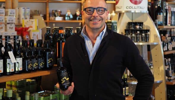 Man with glasses holding a bottle of olive oil in a store filled with various oil products. - Olive Oil Times