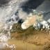 Satellite view showing smoke rising from a wildfire in a landscape with clouds and terrain visible. - Olive Oil Times