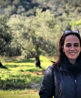A woman with dark hair and sunglasses standing in an olive grove with trees in the background. - Olive Oil Times