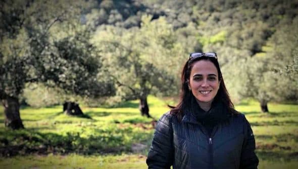 A woman with dark hair and sunglasses standing in an olive grove with trees in the background. - Olive Oil Times