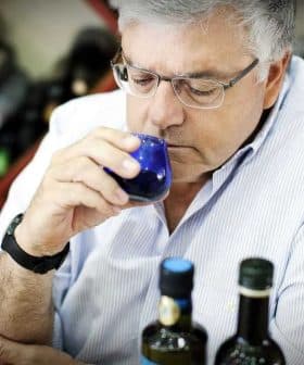 A man with glasses smelling olive oil from a small blue cup while seated at a table with bottles. - Olive Oil Times