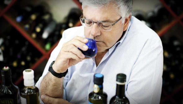 A man with glasses smelling olive oil from a small blue cup while seated at a table with bottles. - Olive Oil Times