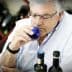 A man with glasses smelling olive oil from a small blue cup while seated at a table with bottles. - Olive Oil Times
