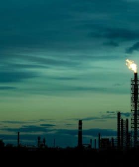 Flare stack emitting flame against a dusk sky with clouds and silhouettes of industrial structures. - Olive Oil Times