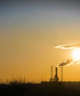 Silhouette of industrial buildings and smokestacks against a sunset sky with a crane in the foreground. - Olive Oil Times