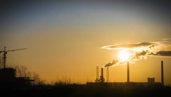 Silhouette of industrial buildings and smokestacks against a sunset sky with a crane in the foreground. - Olive Oil Times