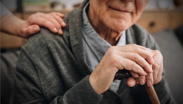 Elderly man with gray hair holding a cane, wearing a gray cardigan, with a hand on his shoulder. - Olive Oil Times