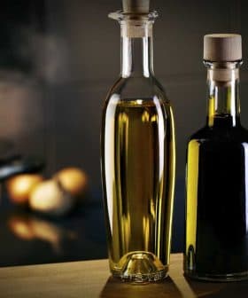 Two glass bottles containing olive oil, one with a light golden liquid and the other with a dark liquid, placed on a wooden surface. - Olive Oil Times