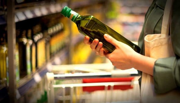 Individual holding a green bottle of olive oil while shopping in a grocery store aisle. - Olive Oil Times