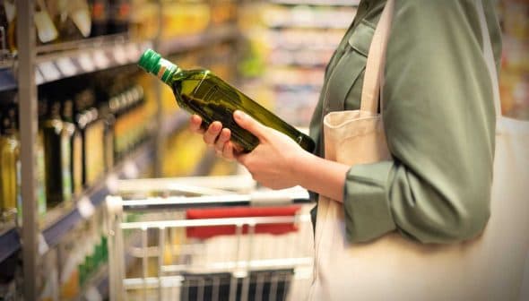 Individual holding a green bottle of olive oil while shopping in a grocery store aisle. - Olive Oil Times