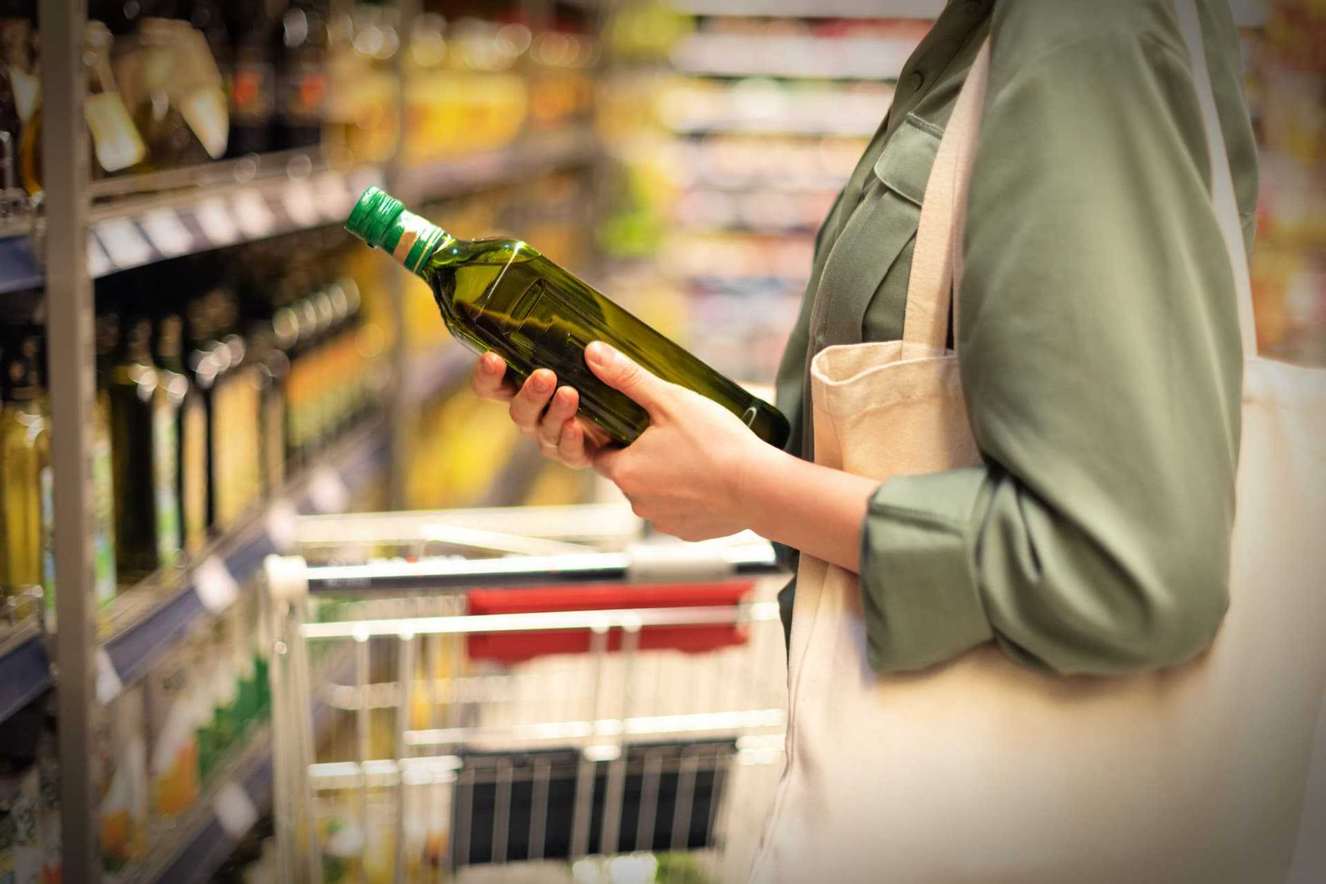 Individual holding a green bottle of olive oil while shopping in a grocery store aisle. - Olive Oil Times