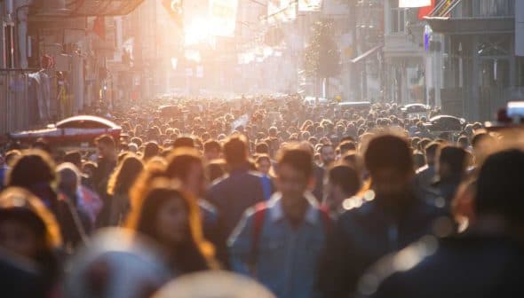 A large crowd of people walking in a busy urban street during sunset with sunlight filtering through. - Olive Oil Times