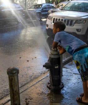 A child in swim trunks leaning against a fire hydrant with water spraying out onto the street. - Olive Oil Times