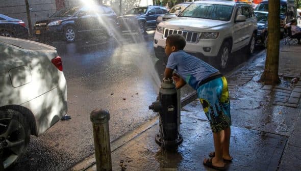 A child in swim trunks leaning against a fire hydrant with water spraying out onto the street. - Olive Oil Times