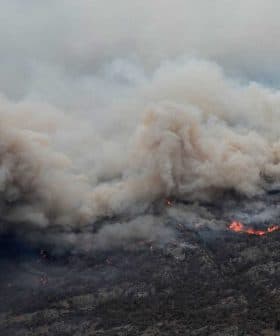Thick smoke billowing from a wildfire with visible flames in a mountainous region. - Olive Oil Times