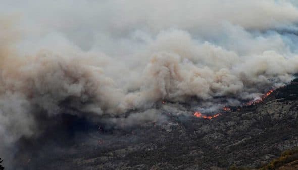 Thick smoke billowing from a wildfire with visible flames in a mountainous region. - Olive Oil Times