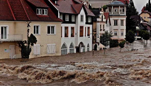 Residential buildings partially submerged in floodwaters with strong currents visible. - Olive Oil Times