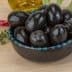 A bowl filled with black olives on a wooden surface with herbs and spices nearby. - Olive Oil Times