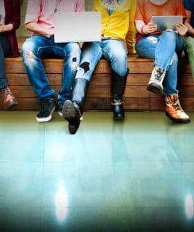 A group of individuals sitting on a bench, each using various electronic devices such as laptops and tablets. - Olive Oil Times
